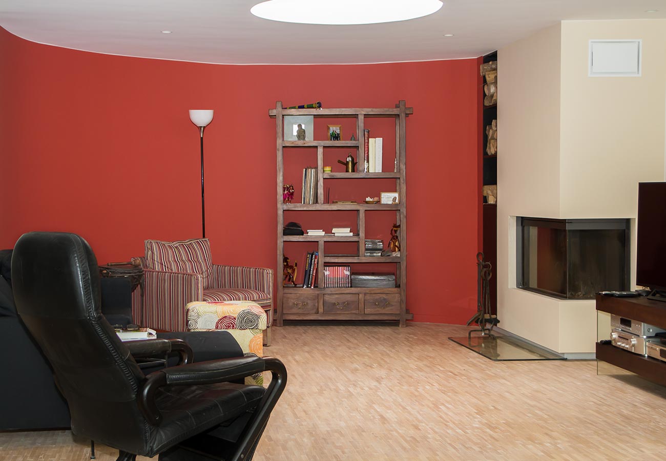 Woonkamer met rood geverfde accentmuur door In't Kleur, een houten boekenkast en een moderne open haard.
