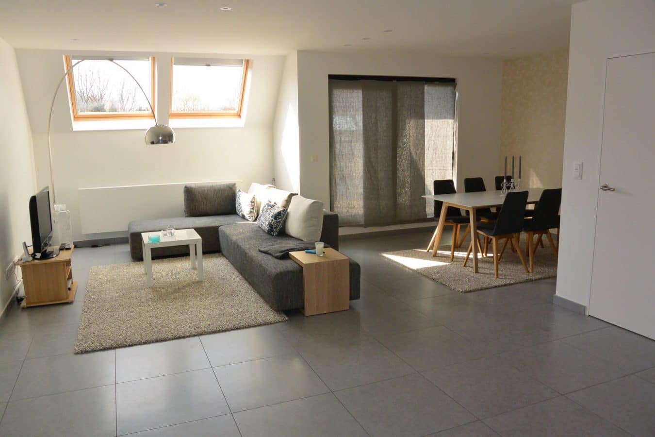 Woonkamer met veel lichtinval dankzij dakramen en een strakke indeling met grijze tegels en witte muren door In't Kleur in Gentbrugge.