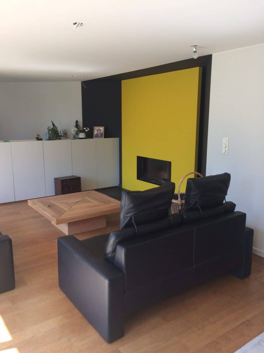Modern ingerichte woonkamer met zwarte leren zetels en een gele wand rond de haard, schilderwerken door In't Kleur in Waregem