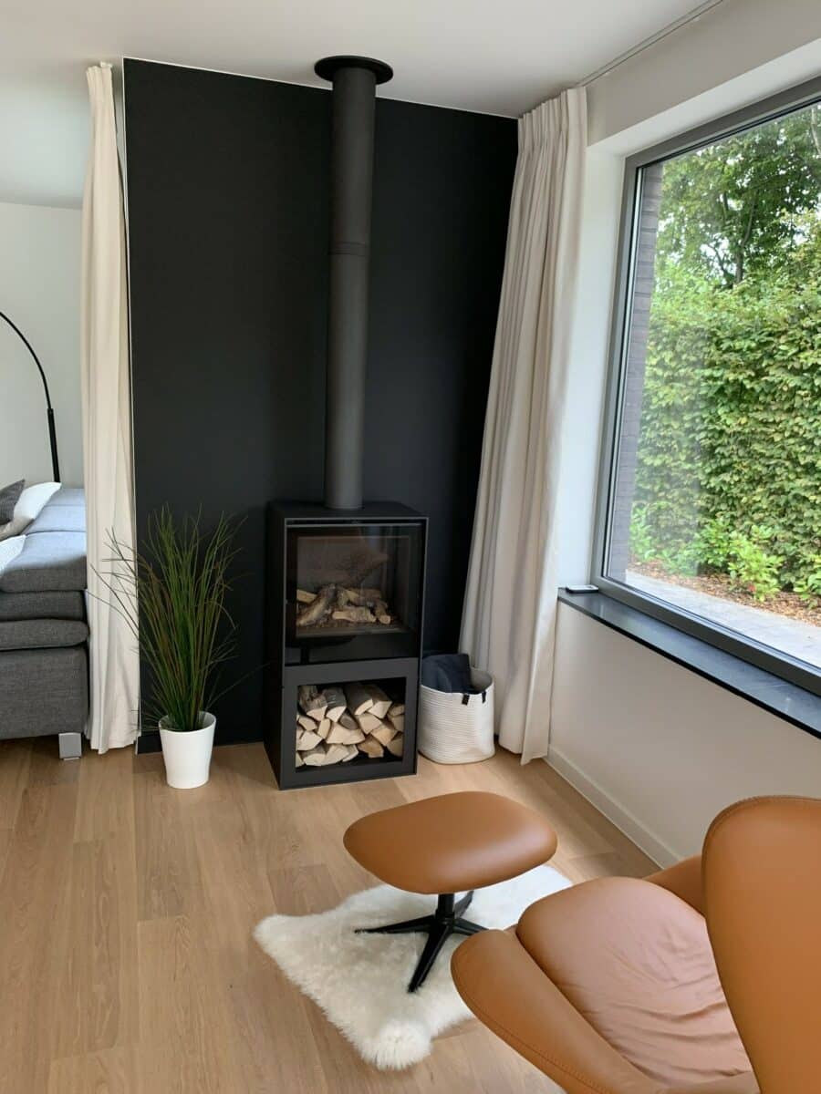 Zithoekje met leren fauteuil, moderne houtkachel voor een zwarte wand door In't Kleur in Moerbeke.