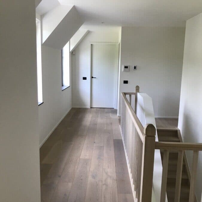 Heldere gang met houten balustrade, witte muren en deuren bij een project in Sint-Martens-Latem.