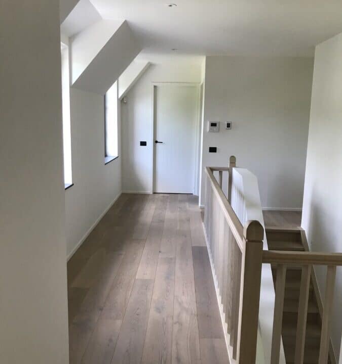 Heldere gang met houten balustrade, witte muren en deuren bij een project in Sint-Martens-Latem.