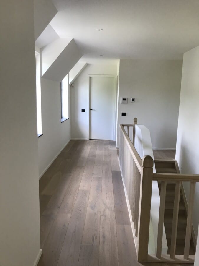 Heldere gang met houten balustrade, witte muren en deuren bij een project in Sint-Martens-Latem.