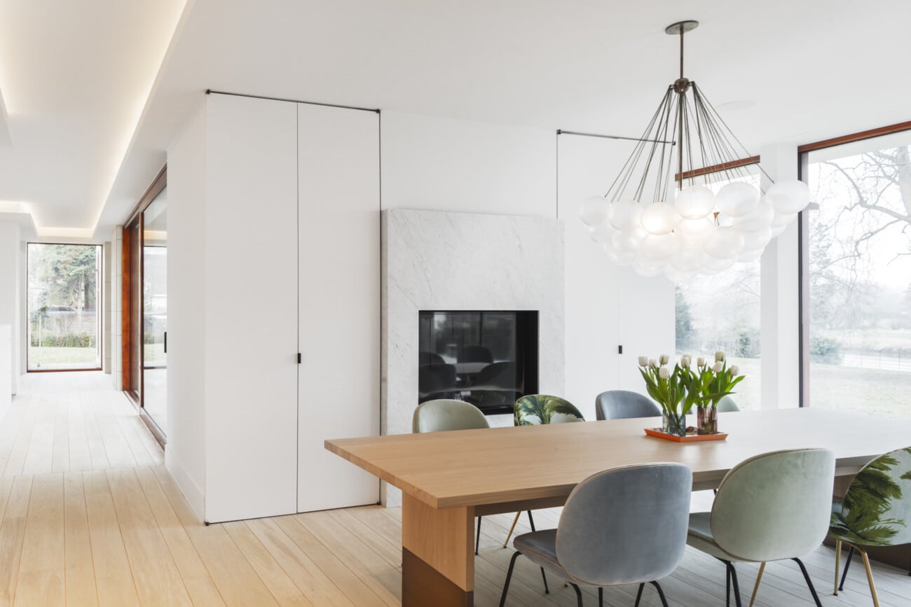 Moderne eetkamer met strakke afwerking - witte muren, naadloos schilderwerk, houten eettafel en grijze stoelen onder een bolvormige kroonluchter, natuurlijk licht door grote ramen met zicht op groene omgeving. Schilderwerken door In't Kleur.