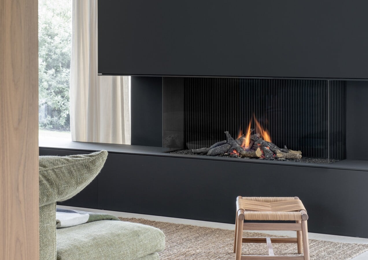 Gezellig interieur met haard en zitruimte - zwart geschilderde haard, brandend vuur, groen kussen op stoel en houten kruk in woonkamer met naadloos schilderwerk door In't Kleur.