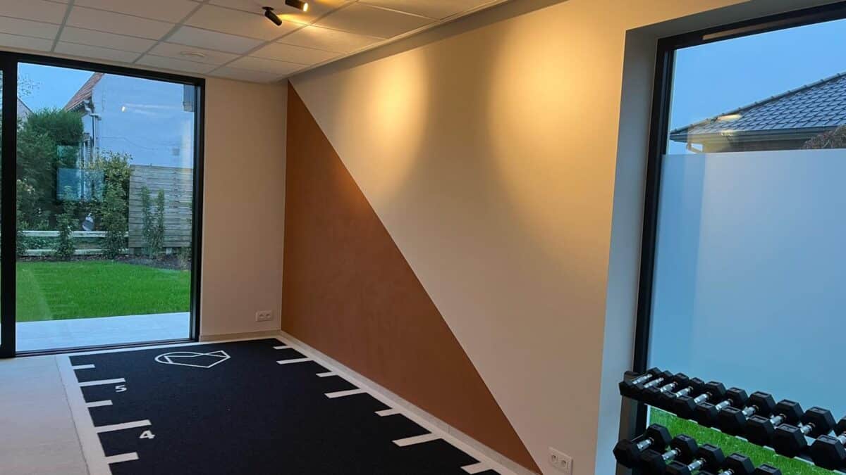 Lievegem kinépraktijk schilderwerken. We zien een accentmuur die half bruin en half gebroken wit geschilderd is, met een schuine streep om beide delen te scheiden. Op de grond ligt een zwarte sportvloer met witte markeringen.