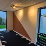 Lievegem kinépraktijk schilderwerken. We zien een accentmuur die half bruin en half gebroken wit geschilderd is, met een schuine streep om beide delen te scheiden. Op de grond ligt een zwarte sportvloer met witte markeringen.