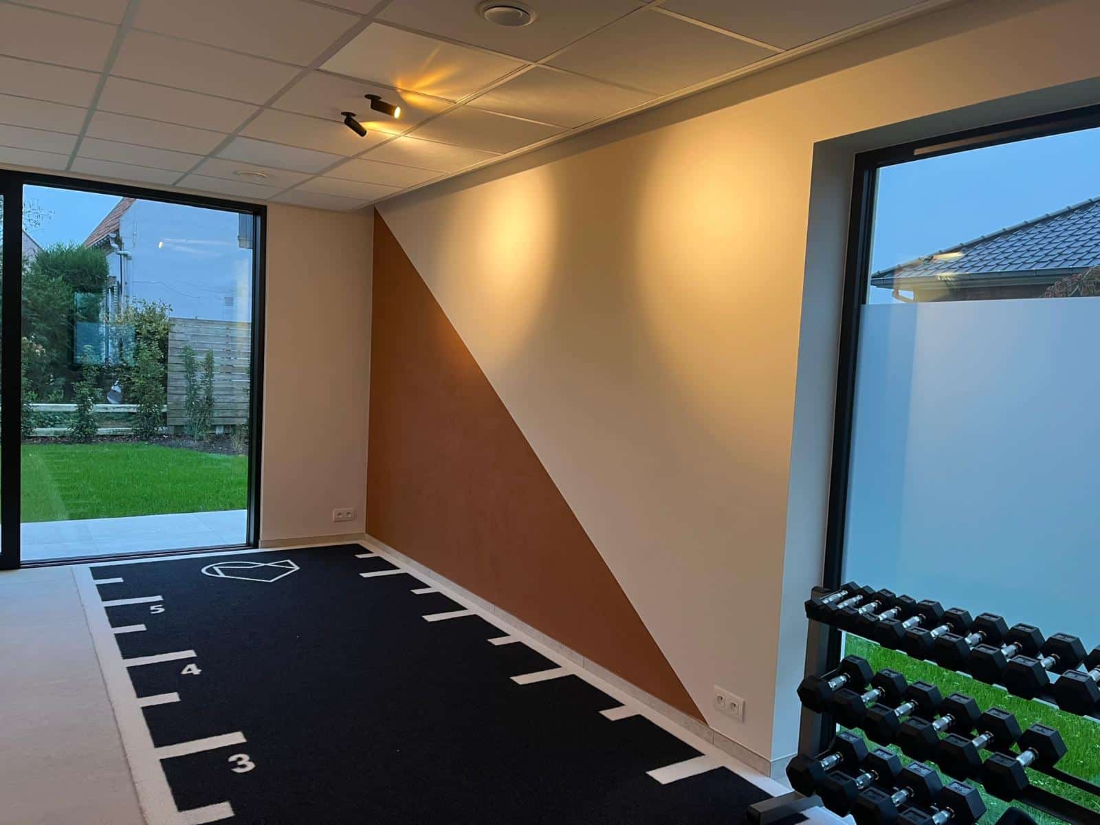 Lievegem kinépraktijk schilderwerken. We zien een accentmuur die half bruin en half gebroken wit geschilderd is, met een schuine streep om beide delen te scheiden. Op de grond ligt een zwarte sportvloer met witte markeringen.