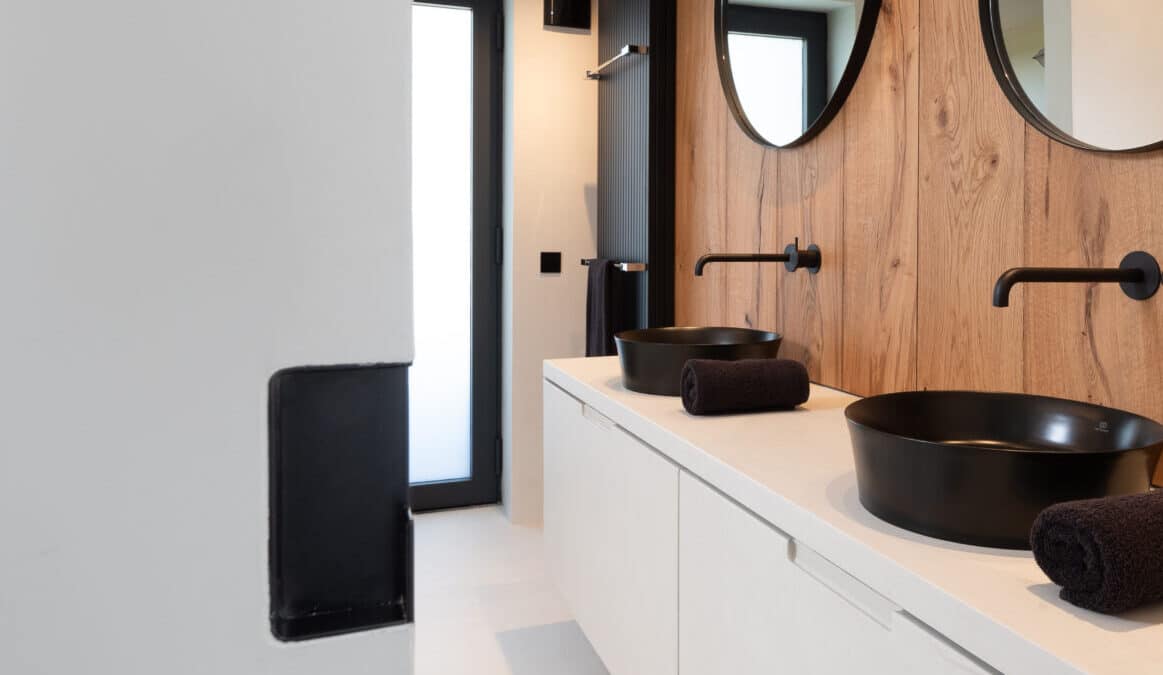 Gerenoveerde badkamer, ontwerp van Herman Interieur, uitgevoerd door In't Kleur en In't Wit. We zien veel hout, beige mortex en zwarte kranen en lavabo's en radiatoren en een zwarte wasmand. En spiegels.