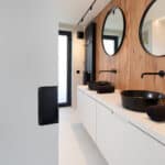 Gerenoveerde badkamer, ontwerp van Herman Interieur, uitgevoerd door In't Kleur en In't Wit. We zien veel hout, beige mortex en zwarte kranen en lavabo's en radiatoren en een zwarte wasmand. En spiegels.