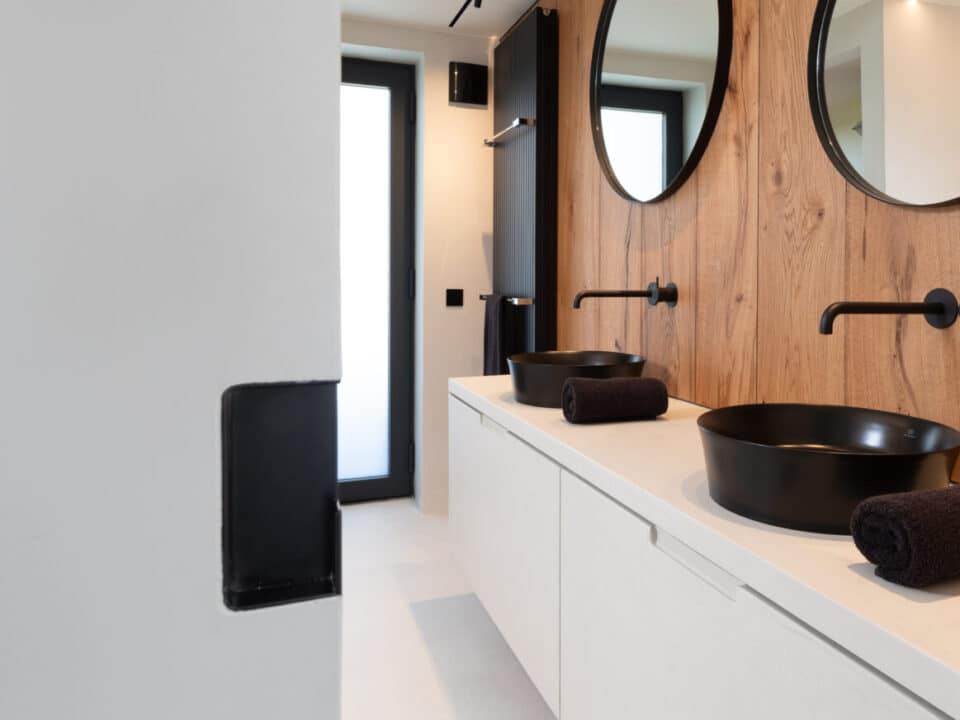 Gerenoveerde badkamer, ontwerp van Herman Interieur, uitgevoerd door In't Kleur en In't Wit. We zien veel hout, beige mortex en zwarte kranen en lavabo's en radiatoren en een zwarte wasmand. En spiegels.