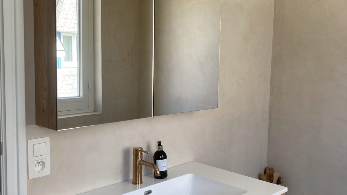 Totale badkamerrenovatie in De Haan door In't Kleur en In't Wit. Op de foto zien we een spiegel met badkamermeubel en beige-lichtbruine Mortex op de muren + een hoekje van het bad.