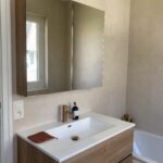 Totale badkamerrenovatie in De Haan door In't Kleur en In't Wit. Op de foto zien we een spiegel met badkamermeubel en beige-lichtbruine Mortex op de muren + een hoekje van het bad.