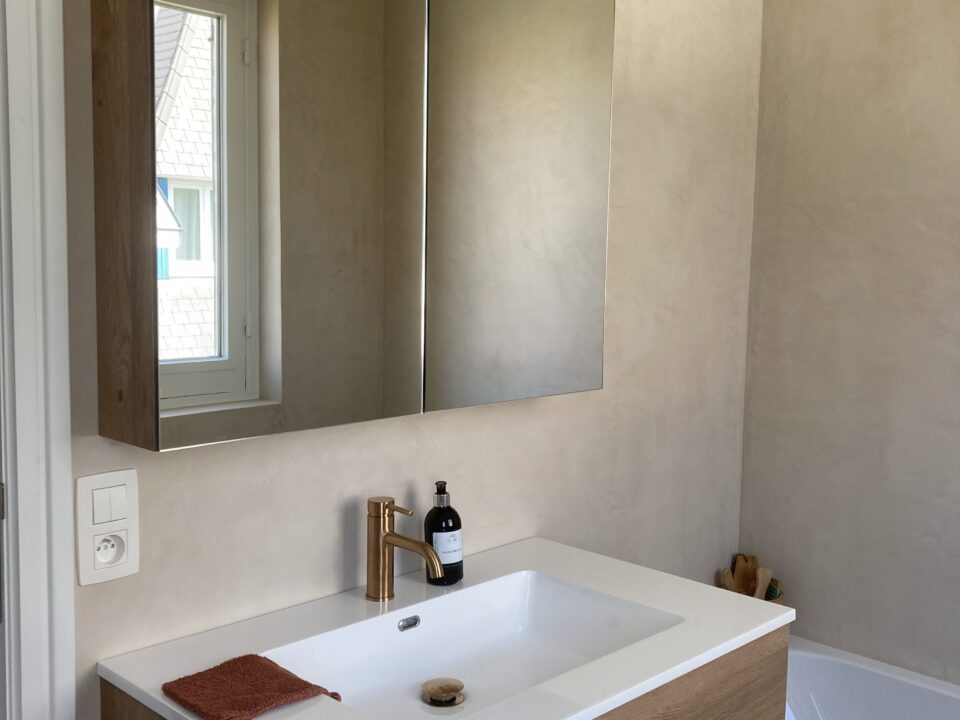 Totale badkamerrenovatie in De Haan door In't Kleur en In't Wit. Op de foto zien we een spiegel met badkamermeubel en beige-lichtbruine Mortex op de muren + een hoekje van het bad.