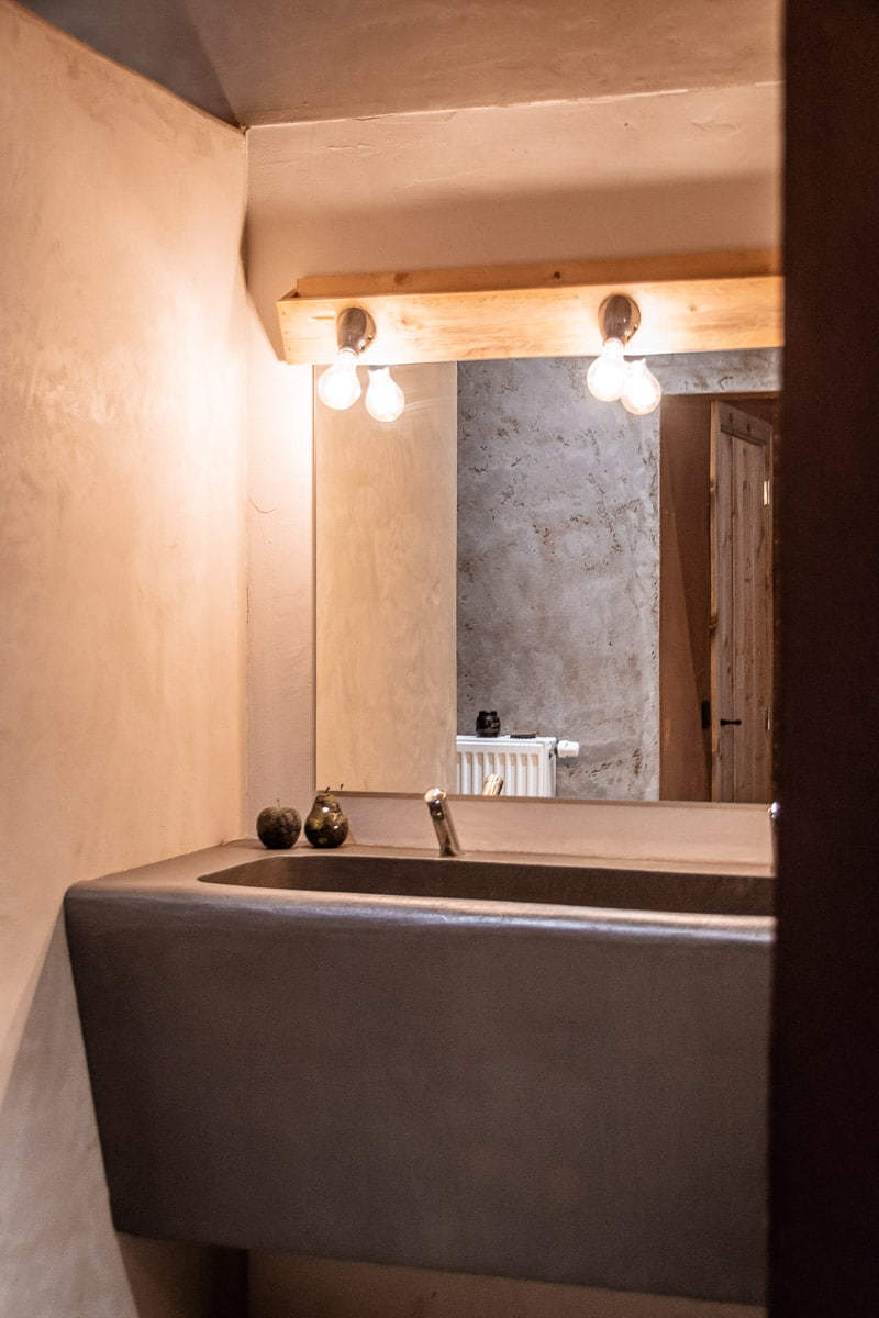 Sfeervolle badkamer met betonnen wastafel, warme verlichting op houten balk en decoratief gepleisterde muren